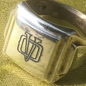 Könnte beinhalten: Nahaufnahme eines silbernen Siegelrings. Der Ring hat eine rechteckige Oberfläche mit einem eingravierten Monogramm-Design. Der Ring besteht aus einem glänzenden, reflektierenden Metall, und das Monogramm hat einen dunkleren Ton, wodurch ein Kontrast entsteht. Ein klassisches Schmuckstück.