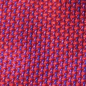 May include: Close-up of a red woven fabric with a textured pattern. The weave reveals small areas of blue and purple threads, creating a subtle contrast within the red background. The fabric has a tight, uniform structure.