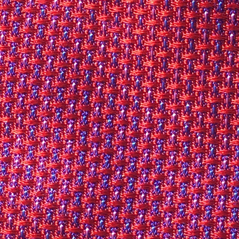 May include: Close-up of a red woven fabric with a textured pattern. The weave reveals small areas of blue and purple threads, creating a subtle contrast within the red background. The fabric has a tight, uniform structure.