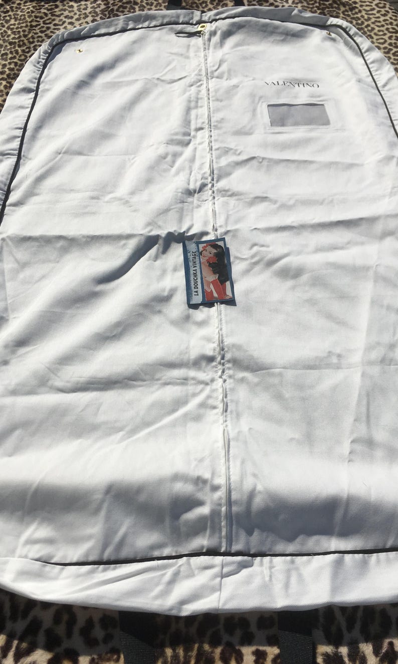 May include: A white garment bag with a black trim and a zipper closure. The bag has the brand name "VALENTINO" printed on it. A tag with the text "LA DOUCHKA VINTAGE" and an illustration is attached to the bag.