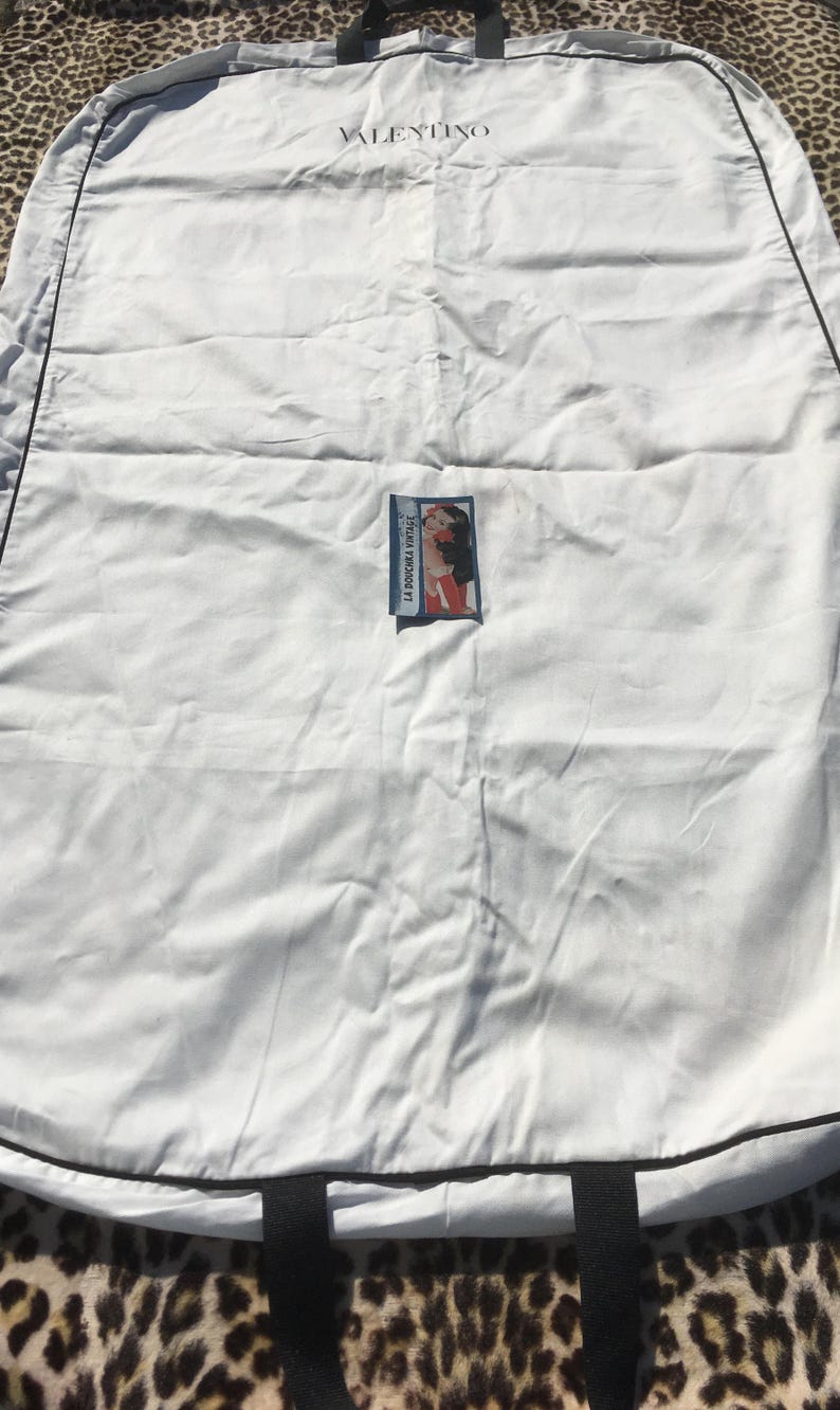 May include: A white Valentino garment bag with black trim and handles. The brand name is printed across the top. A small rectangular label with a vintage illustration is attached to the front.
