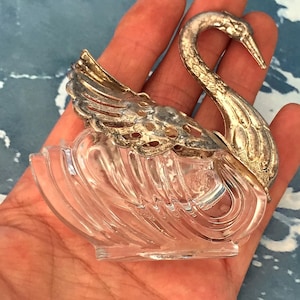 Puede incluir: Una figurita de cisne de vidrio transparente con cabeza y cuello plateados. El cisne tiene un diseño detallado y es una pieza decorativa.