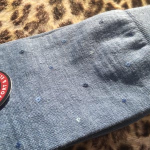 Peut inclure: Gros plan sur une chaussette bleue avec de petits points bleu foncé et bleu clair dispersés. La chaussette a un bord-côte et un aspect doux et texturé. Une partie d'une étiquette circulaire avec du texte rouge et blanc est visible.
