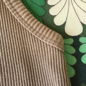 May include: Close-up of a ribbed, tan-colored garment. The fabric has vertical lines and a textured appearance. The neckline is also ribbed. The background features a green and white floral pattern.