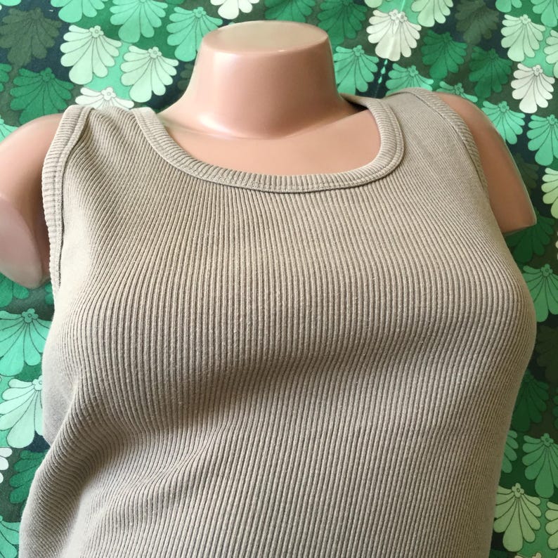 May include: A light olive green ribbed tank top. The sleeveless top has a scoop neckline and is displayed on a mannequin. The fabric has vertical ribbing. The background is a green and white floral pattern.