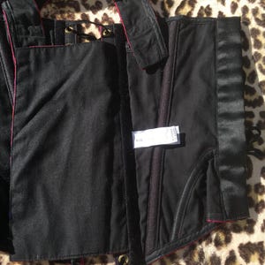 May include: Black corset with red trim and satin accents. The corset features a structured design with boning, eyelets, and a label with size information. The garment is made of a matte fabric with a satin panel and is displayed on a leopard print surface.