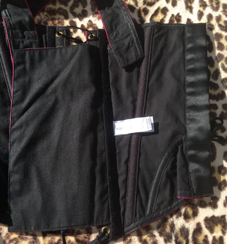 May include: Black corset with red trim and satin accents. The corset features a structured design with boning, eyelets, and a label with size information. The garment is made of a matte fabric with a satin panel and is displayed on a leopard print surface.