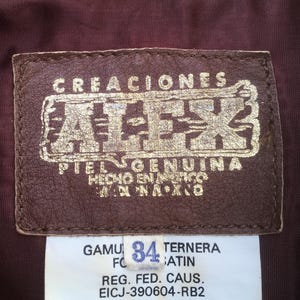 Puede incluir: Primer plano de una etiqueta de cuero marr&oacute;n con letras doradas: "CREACIONES ATEK PIEL GENUINA HECHO EN MEXICO." Debajo, una etiqueta blanca muestra "GAMU 34 TERNERA" y otro texto. El fondo es de tela burdeos.