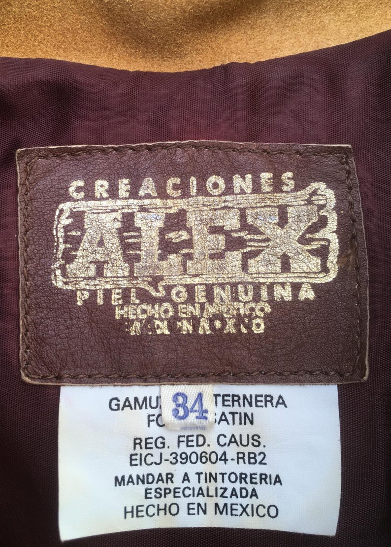 Puede incluir: Primer plano de una etiqueta de cuero marr&oacute;n con letras doradas: "CREACIONES ATEK PIEL GENUINA HECHO EN MEXICO." Debajo, una etiqueta blanca muestra "GAMU 34 TERNERA" y otro texto. El fondo es de tela burdeos.