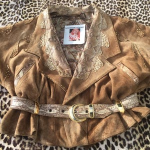 Könnte beinhalten: Braune Wildlederjacke mit Schlangenmuster-Kragen, Revers und Gürtel. Die Jacke hat goldfarbene Nieten und eine große goldene Schnalle am Gürtel. Das Etikett lautet "LA BOUCHKA VINTAGE".