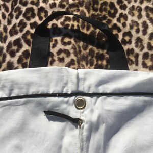 May include: A white tote bag with a black handle and trim. The bag has a drawstring closure with a black cord and a metal grommet. The background is a leopard print pattern.