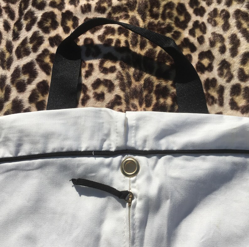 May include: A white tote bag with a black handle and trim. The bag has a drawstring closure with a black cord and a metal grommet. The background is a leopard print pattern.