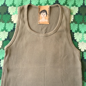 May include: Olive green ribbed tank top with a scoop neck. A small tag with the text "LA DOUCHKA VINTAGE" and a drawing of a woman is attached to the neckline. The tank top is made of a soft, ribbed material.