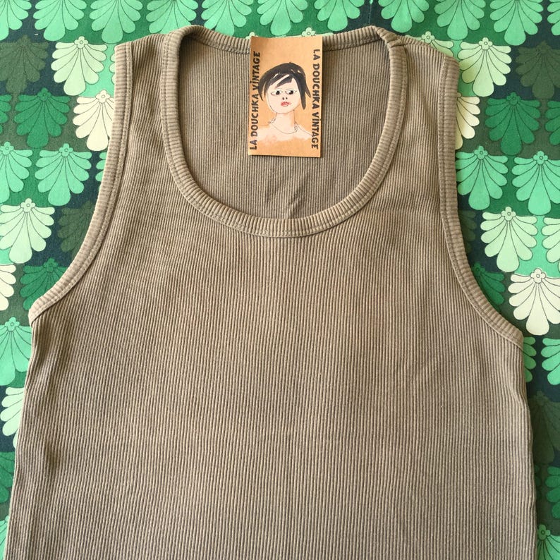 May include: Olive green ribbed tank top with a scoop neck. A small tag with the text "LA DOUCHKA VINTAGE" and a drawing of a woman is attached to the neckline. The tank top is made of a soft, ribbed material.