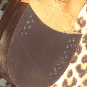 May include: Close-up of a brown leather shoe sole with metal studs. The sole is a dark brown color and has a curved shape. The studs are small and silver, arranged in rows along the edge of the sole. The shoe's interior is visible, with the word "FORME" stamped on the leather.