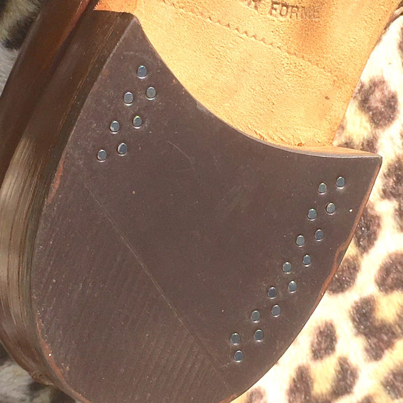 May include: Close-up of a brown leather shoe sole with metal studs. The sole is a dark brown color and has a curved shape. The studs are small and silver, arranged in rows along the edge of the sole. The shoe's interior is visible, with the word "FORME" stamped on the leather.
