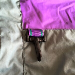 May include: A close-up of a black plastic hook with a purple and green striped strap attached to a gray and purple fabric.
