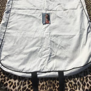 May include: A white garment bag with black trim and handles. A small label with a woman's image is attached to the bag. The bag is lying on a leopard print surface.