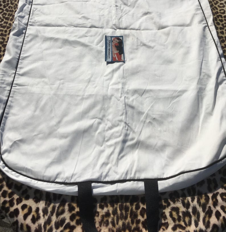 May include: A white garment bag with black trim and handles. A small label with a woman's image is attached to the bag. The bag is lying on a leopard print surface.
