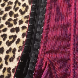 May include: Close-up of a red and purple textured corset with black trim and metal hook closures. The corset is partially open, revealing the interior lining. The background features a leopard print pattern.