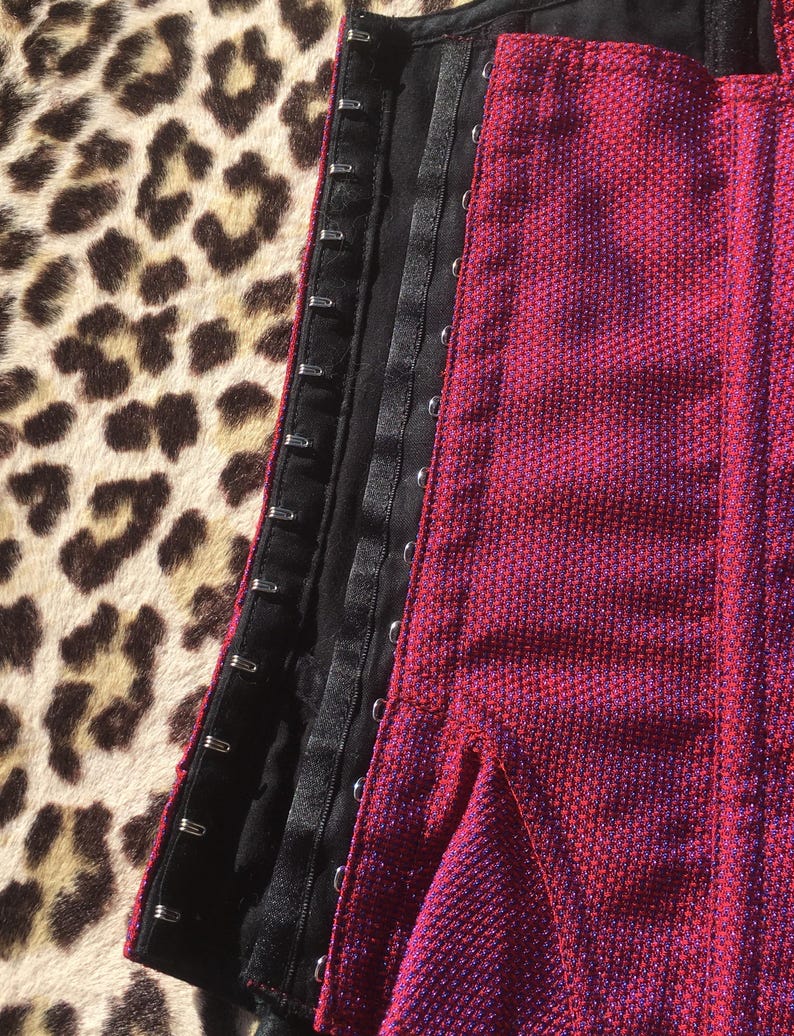 May include: Close-up of a red and purple textured corset with black trim and metal hook closures. The corset is partially open, revealing the interior lining. The background features a leopard print pattern.