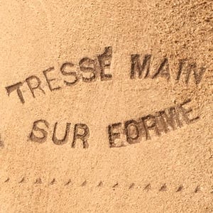 May include: Close-up of a tan leather surface with the embossed text "TRESSE MAIN SUR FORME". The text is in a dark brown color. A dotted line runs along the bottom of the image.