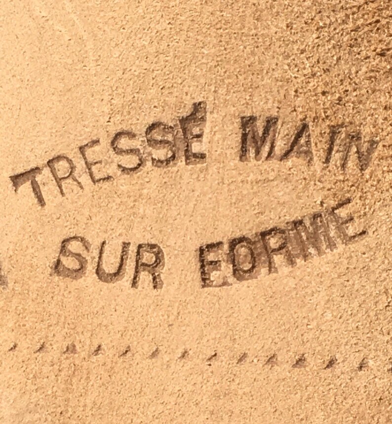 May include: Close-up of a tan leather surface with the embossed text "TRESSE MAIN SUR FORME". The text is in a dark brown color. A dotted line runs along the bottom of the image.