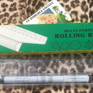 Puede incluir: Una caja verde y amarilla con la inscripción "Multi-Purpose Rolling Ruler" y una regla rodante blanca y gris dentro de una funda de plástico transparente. La caja presenta formas geométricas y un anuncio vintage.