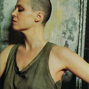 May include: A side profile portrait of a person with a shaved head, wearing a green tank top. The person is looking to the left, with a neutral expression. The background is a textured wall with faded colors.