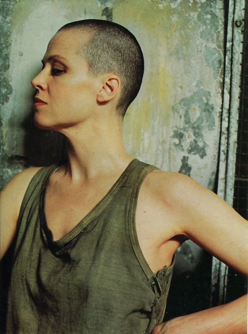 May include: A side profile portrait of a person with a shaved head, wearing a green tank top. The person is looking to the left, with a neutral expression. The background is a textured wall with faded colors.