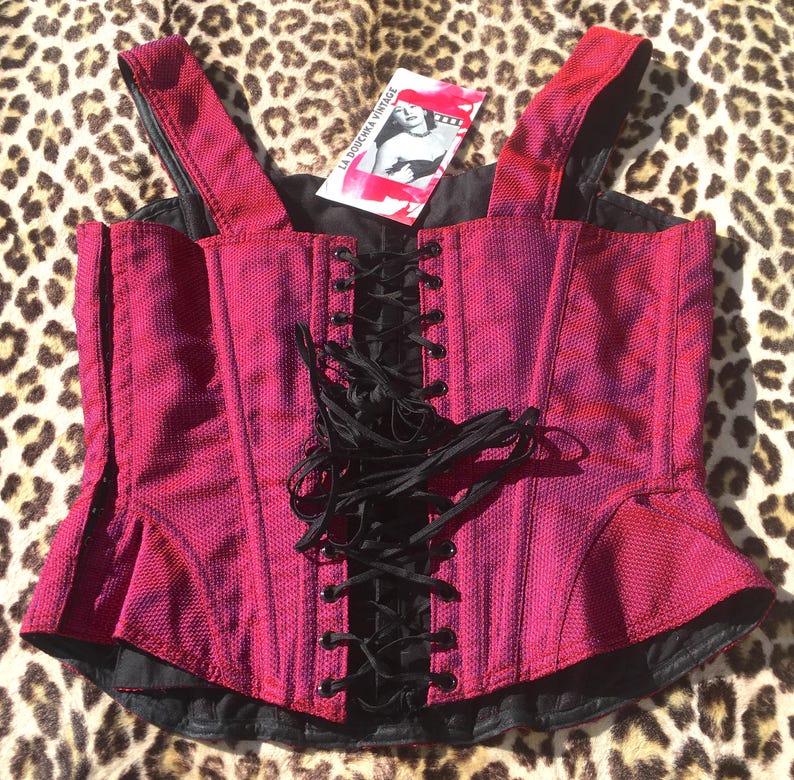 May include: A magenta corset top with black lacing and straps. The top has a textured fabric and a peplum-style hem. A tag with the text "LA DOUCHKA VINTAGE" is attached. The corset is displayed on a leopard print background.