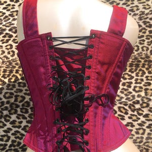 May include: A magenta corset with black lacing, shoulder straps, and a textured fabric. The back of the corset is visible, highlighting the lacing detail. The corset is displayed on a mannequin.
