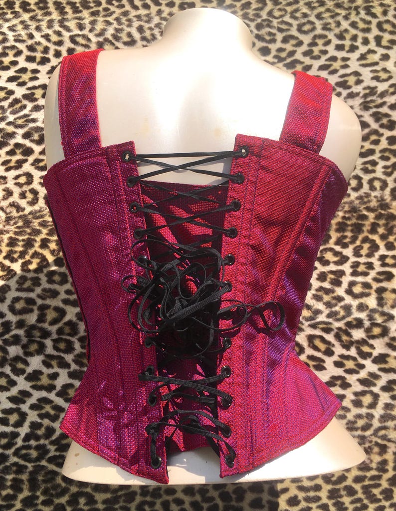 May include: A magenta corset with black lacing, shoulder straps, and a textured fabric. The back of the corset is visible, highlighting the lacing detail. The corset is displayed on a mannequin.