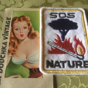 Può includere: Una cartolina vintage con una donna sorpresa con top bianco e gonna rossa, accanto a una toppa bianca con le parole "SOS NATURE" e un grafico di un albero, fuoco e un fiammifero.