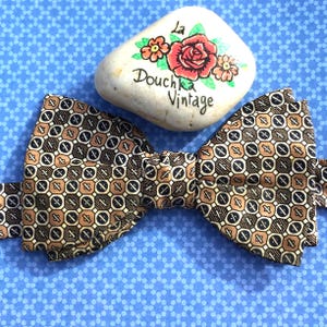 Puede incluir: Una pajarita con un patrón geométrico repetitivo en tonos marrón, negro y blanco. Sobre la pajarita hay una piedra lisa de color claro pintada con una rosa roja, hojas verdes y el texto La Douchka Vintage escrito en tinta negra.