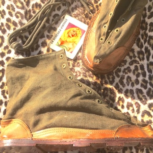 Puede incluir: Un par de botas de tobillo de lona verde y cuero marrón con cordones. Las botas tienen una suela gruesa de goma y están atadas con un cordón verde. Las botas están sobre una tela de estampado de leopardo.