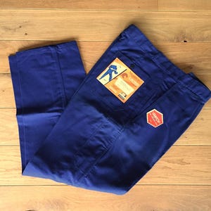Puede incluir: Pantalones de trabajo azul real con cierre de botón y múltiples bolsillos. Los pantalones tienen un bolsillo rectangular en la parte trasera y un bolsillo cuadrado en la parte delantera. Una etiqueta con un gráfico de una persona está adjunta.
