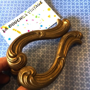 May include: Two ornate gold-toned metal drawer pulls with a decorative scroll design. The pulls are in the shape of an 'S' and have a vintage look.