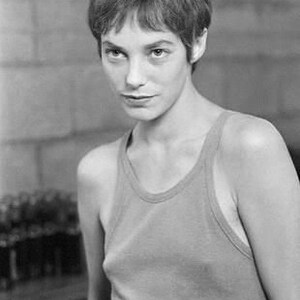 May include: Black and white portrait of a person with short, textured hair and a tank top. The person is looking towards the camera. The background is a textured wall.