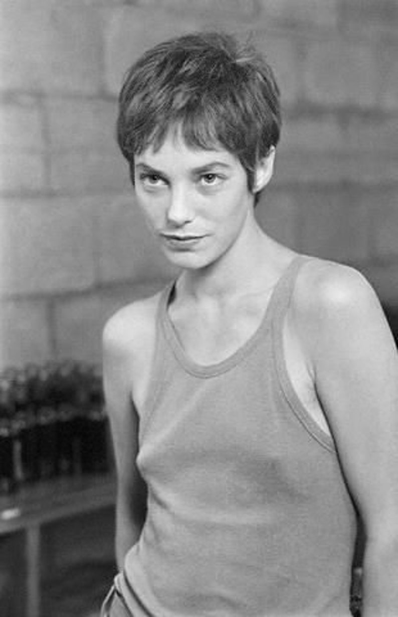 May include: Black and white portrait of a person with short, textured hair and a tank top. The person is looking towards the camera. The background is a textured wall.