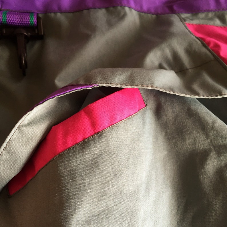 May include: Close-up of a gray jacket with a pink pocket. The jacket has a purple trim.