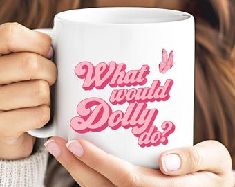 Puede incluir: Una taza de cerámica blanca con el texto rosa "What would Dolly do?" en una fuente de estilo retro. El diseño incluye un pequeño gráfico de una mariposa rosa colocado sobre el texto. La taza tiene un asa estándar y un acabado liso.