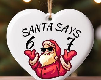 May include: A white heart-shaped Christmas ornament with the text "SANTA SAYS 6 7". The ornament features a cartoon Santa Claus wearing sunglasses and a red suit. The ornament hangs from a brown string.