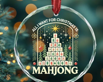 May include: A clear, round Christmas ornament with the text "ALL I WANT FOR CHRISTMAS IS MAHJONG" in a festive design. The ornament hangs from a red ribbon, with a Christmas tree and other ornaments in the background.