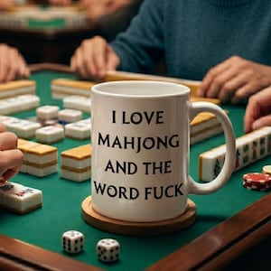 May include: A white ceramic mug with the text "I LOVE MAHJONG AND THE WORD FUCK" sits on a wooden coaster. The mug is on a green table with mahjong tiles and dice. People are playing mahjong in the background.