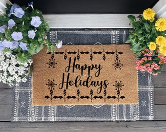 May include: A rectangular coir doormat with the words "Happy Holidays" in black script. The mat is decorated with black ornaments and snowflakes. The mat is on a patterned rug, with potted flowers on either side.