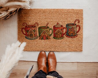 May include: A rectangular doormat with a brown coir surface features four colorful mugs of hot chocolate. Each mug has a gingerbread house design and a candy cane or spoon. The doormat is a festive holiday decoration.