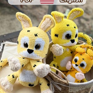 Op de afbeelding: Drie gele gehaakte knuffels met zwarte stippen, witte gezichtsvlekken en grote donkere ogen. De tekst bovenaan luidt Pattern by 2BoyCrochet PDF English. De speeltjes staan in en naast een gevlochten mand op een houten ondergrond.