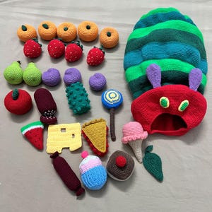 Può includere: Una collezione di oggetti all'uncinetto che include un grande bruco verde e blu con la testa rossa, vari frutti come fragole, arance, pere e prugne, una fetta di anguria, formaggio, torta, una salsiccia, un cupcake, un gelato e un lecca-lecca.