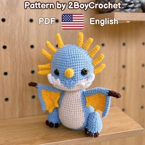 May include: A crocheted blue and yellow dragon with a cream-colored belly. The dragon has black eyes and brown claws. The text "Pattern by 2BoyCrochet" and "PDF English" are visible in the image.